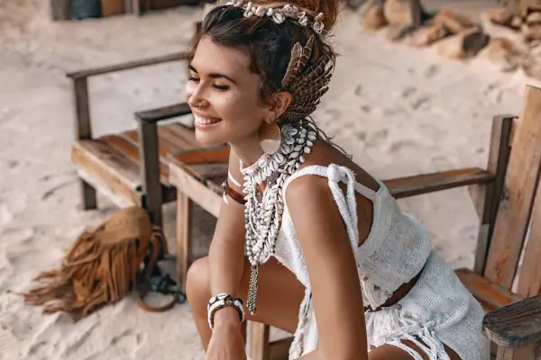 Uśmiechnięta kobieta w stylu boho, ozdobiona muszlami i piórami, siedzi na plaży.